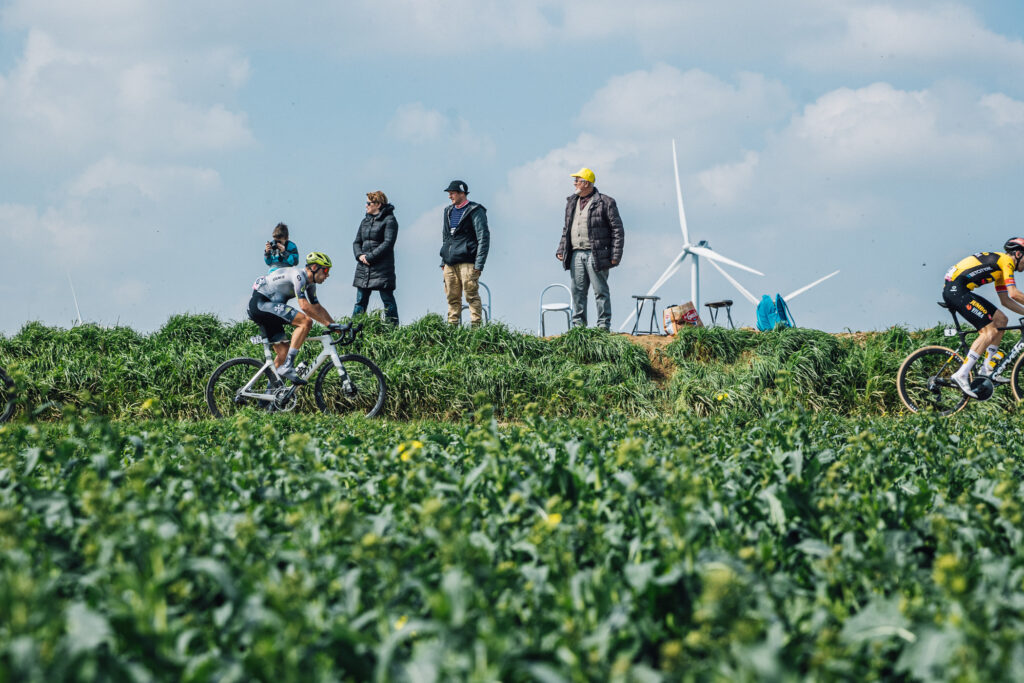 Q36.5 Pro Cycling Team rider cycling through green farmland with wind turbines in the background at the Paris-Roubaix race.