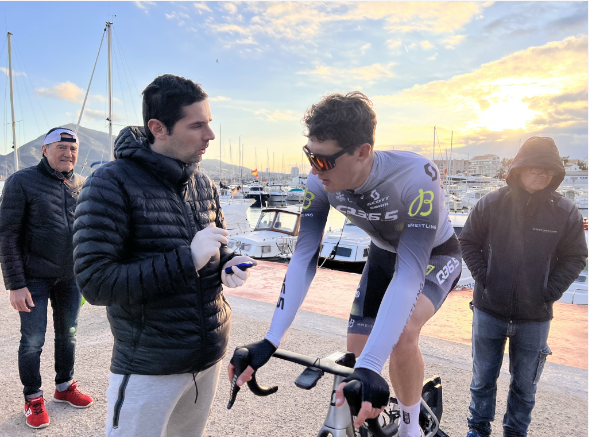 Carles Tur, Head Coach of Q36.5 Pro Cycling Team, advising an athlete cycling on a stationary bike outdoors near a harbour during a training camp.
