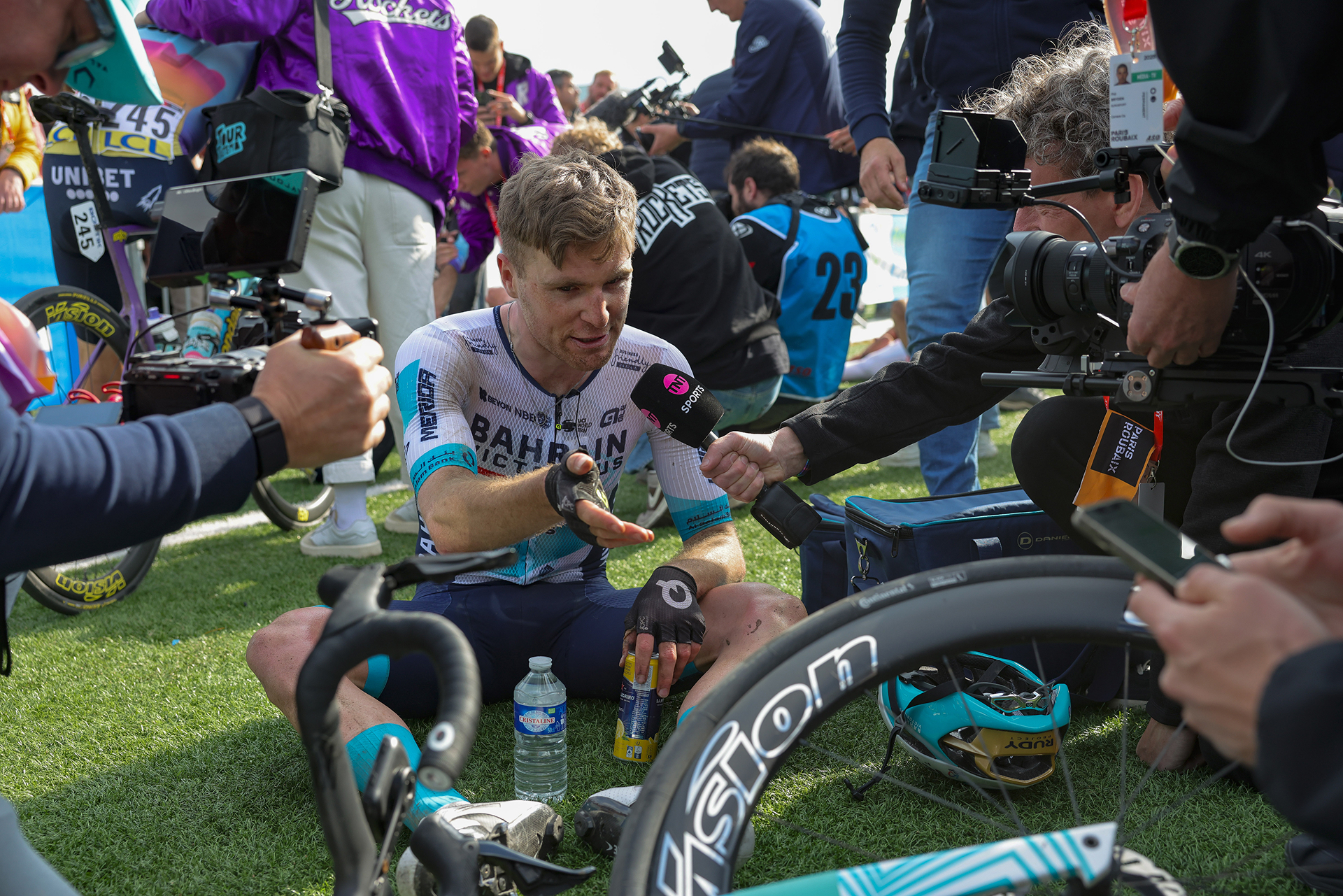 Fred Wright gives a post-race interview after Paris-Roubaix 2025 while sitting on the grass, exhausted and covered in dust, still in his Bahrain-Victorious kit.